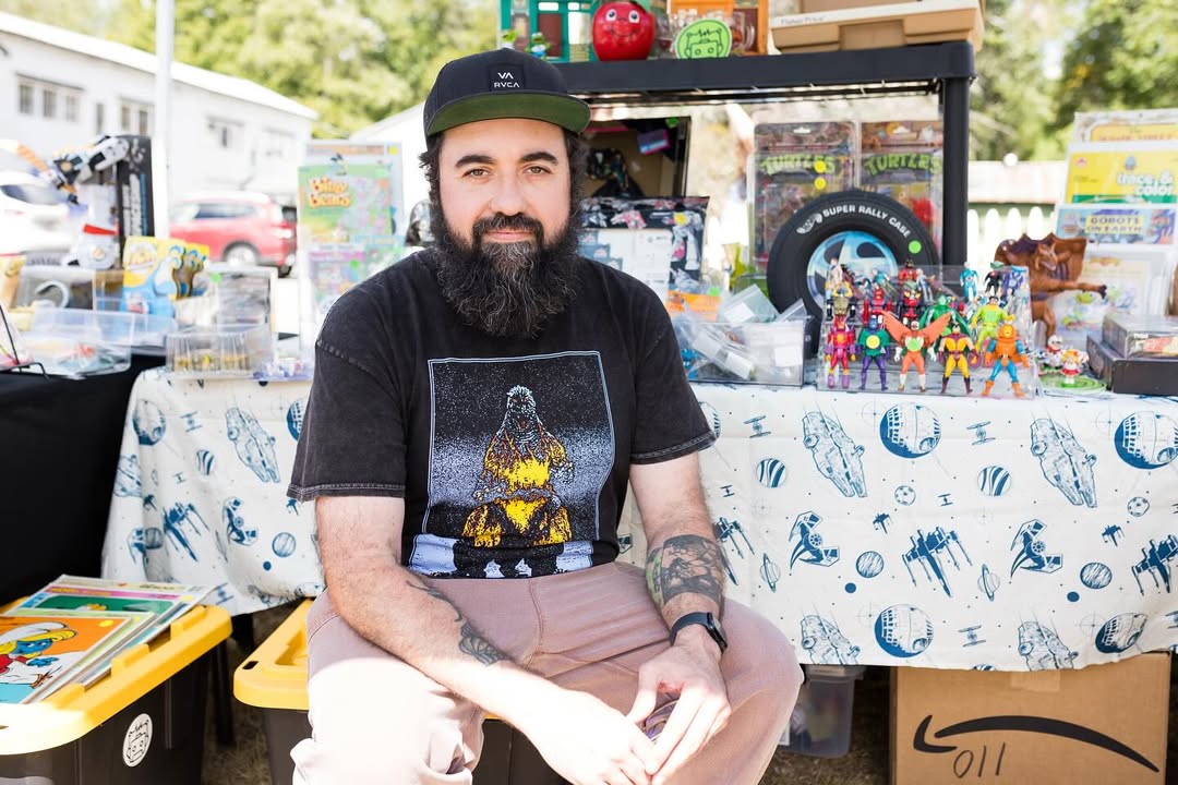 Kneeko, owner of Feast of Ruin, sitting behind a table displayed with retro action figures, VHS tapes, and collectibles at an outdoor toy show.